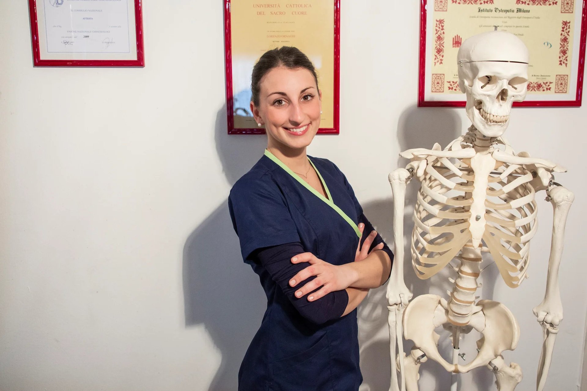 Professionista sanitaria in camice blu scuro che posa accanto a un modello anatomico di scheletro, nello studio con diplomi appesi alla parete
